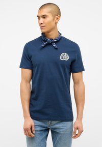 Granatowa bawełniana koszulka z okrągłym dekoltem i białym okrągłym logo. Noszona z wzorzystą niebieską chustą na szyi. Klasyczny krój.