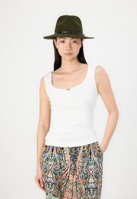Top de tirantes blanco acanalado con un escote redondeado y un acento decorativo en el centro, combinado con pantalones coloridos y estampados y un sombrero verde oscuro.