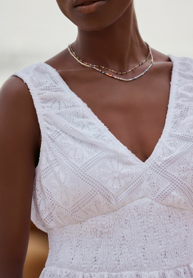 Robe blanche en tissu texturé, ornée de broderies complexes et d'un décolleté en V profond. Portée avec des colliers en perles multicolores superposés.