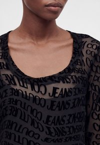 Κοντινό πλάνο ενός ατόμου που φορά ένα διάφανο μαύρο τοπ με μαύρο κείμενο που γράφει «JEANS», «COUTURE» και «VERSACE» σε διάφορες κατευθύνσεις.