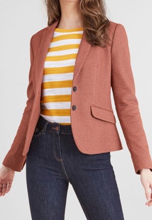 Frau trägt ein rostfarbenes Blazer über einem weiß und senfgelb gestreiften Hemd und dunkelblaue, hoch taillierte Jeans.