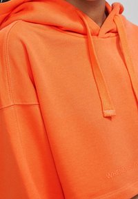 Orange hoodie med en texturerad yta, med en dragsko-hood och broderad text vid nederkanten. Enkelt och modernt design.