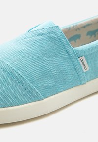 Chaussure slip-on en toile bleu clair avec semelle en caoutchouc blanche et petite étiquette en tissu indiquant "184/200" sur le côté.