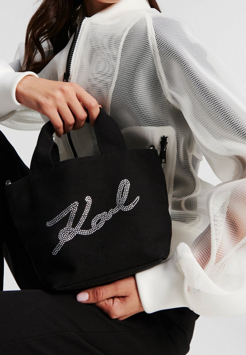 Una donna con una giacca in rete bianca tiene in mano una piccola borsa a tracolla nera con il testo "Kool" in strass sul davanti.