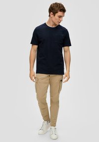 Marineblauwe kortekogel-shirt met een zak, gecombineerd met beige cargobroeken met zijzakken, gedragen met witte sneakers. Glad textuur.