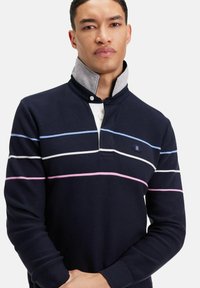 Pull bleu marine foncé à col montant, avec des accents gris. Rayures horizontales en bleu clair, blanc et rose. Poignets côtelés.