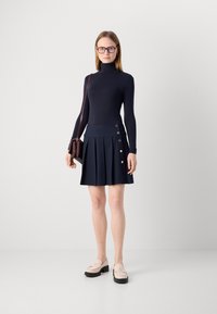 Námořnický rolák s dlouhým rukávem v kombinaci s plisé sukni v námořnické barvě, která má zlaté knoflíkové detaily. Celý outfit doplňují světlé mokasíny.