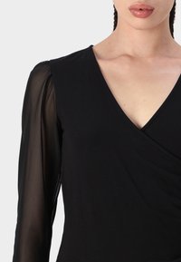 Top nero con scollatura a V. Maniche corte trasparenti con finitura testurizzata. Tessuto del corpo liscio, con uno stile avvolgente nella parte anteriore.