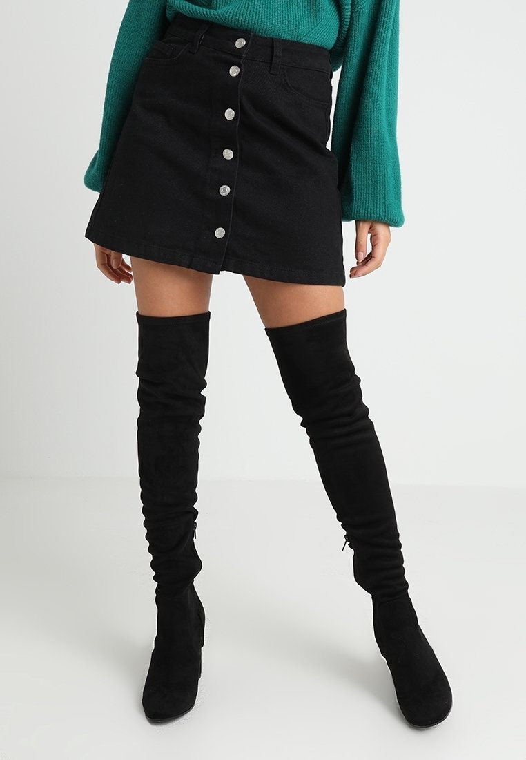 Femme portant une mini-jupe noire à boutons devant, un pull à manches longues bleu sarcelle et des bottes hautes en daim noir, debout sur un fond uni.