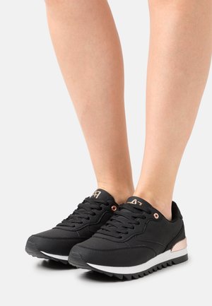 Pieds portant des baskets noires texturées à lacets avec des semelles blanches et des accents en or rose, posés sur une surface gris clair.