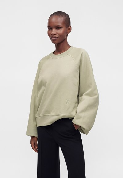 Personne debout devant un fond uni, portant un sweat-shirt ample vert clair et un pantalon noir, une main dans une poche.
