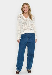Cremefarbener gestrickter Pullover mit V-Ausschnitt, versehen mit einem Spitzenmuster, kombiniert mit blauen weit geschnittenen Denim-Jeans und braunen Slippern.