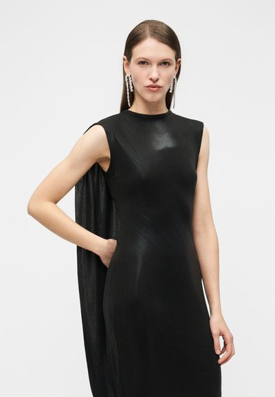 Nő, aki ujjatlan fekete ruhát visel köpenyrésszel és hosszú, csillogó kristályfüggő fülbevalókkal, egyik kezét csípőjén tartva pózol.