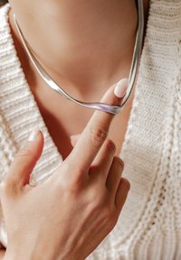 Collana in argento con un design flessibile, che si adagia delicatamente attorno al collo. Una mano con uno smalto neutro tiene la collana, mettendo in evidenza la sua texture.