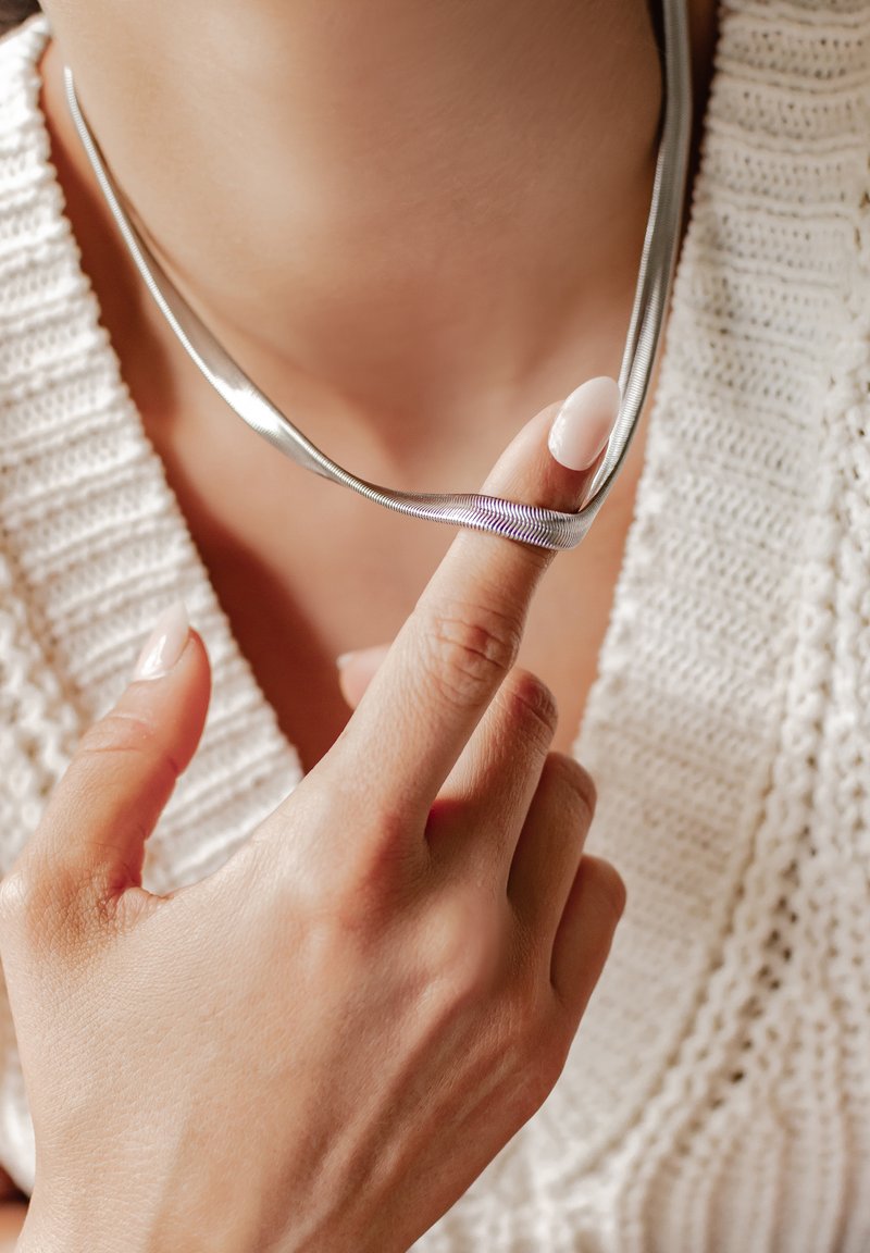 Collana in argento con un design flessibile, che si adagia delicatamente attorno al collo. Una mano con uno smalto neutro tiene la collana, mettendo in evidenza la sua texture.