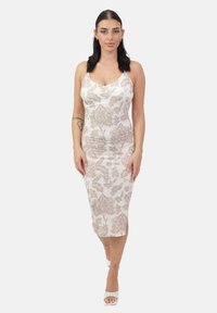 Donna con capelli scuri che indossa un vestito midi floreale bianco e beige e tacchi bianchi, in piedi contro uno sfondo chiaro e semplice.