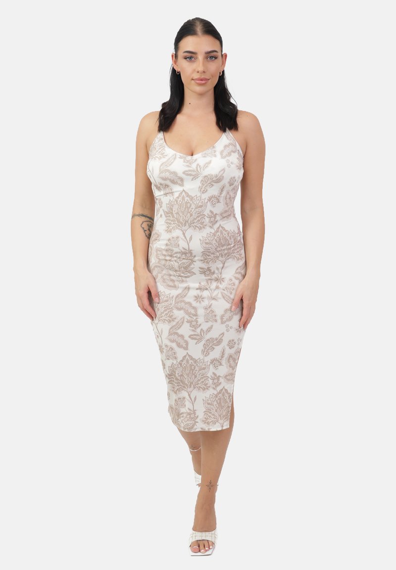 Donna con capelli scuri che indossa un vestito midi floreale bianco e beige e tacchi bianchi, in piedi contro uno sfondo chiaro e semplice.