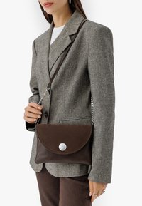 Borsa in pelle marrone con patta pieghevole, hardware in argento e tracolla a catena, esposta su un blazer grigio e pantaloni marroni.