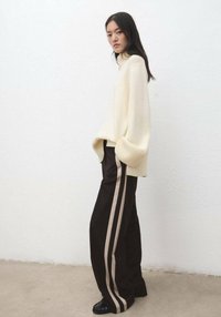 Pull côtelé crème avec manches amples associé à un pantalon noir avec des rayures latérales marron clair ; le mannequin se tient contre un mur blanc.