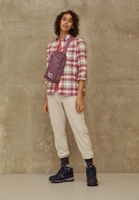 Lila axelremsväska med två bruna detaljer, som bärs över en rosa skjorta och en rutiga skjorta, i kombination med beige cargobyxor och svarta vandringsskor.