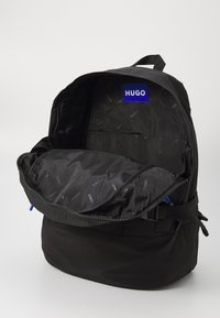 Sac à dos noir avec une ouverture zippée, présentant un extérieur texturé et un intérieur doublé affichant le logo "HUGO" en bleu.