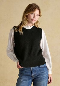 Junge Frau trägt einen schwarzen ärmellosen Pullover über einer weißen Bluse mit Rüschenkragen, kombiniert mit blauen Jeans, vor beigem Hintergrund.