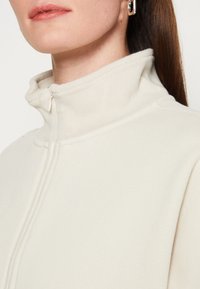 Forro polar crema con cremallera y cuello alto. Presenta textura acanalada y costuras visibles a lo largo de las uniones. Apariencia sutil y de tela suave.