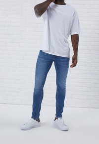Vit oversized t-shirt i kombination med blå skinny jeans och vita sneakers, mot en tegelvägg som bakgrund. Enkelt, avslappnat design.