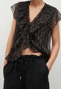 Blouse noire transparente à motif floral, col et manches à volants, longueur courte. Associée à un pantalon noir ample avec une ceinture élastique.