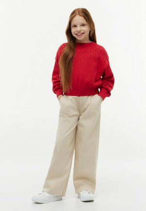 Lächelndes Mädchen mit langen roten Haaren trägt einen roten Pullover, beige weite Hosen und weiße Turnschuhe, steht mit den Händen in den Taschen.