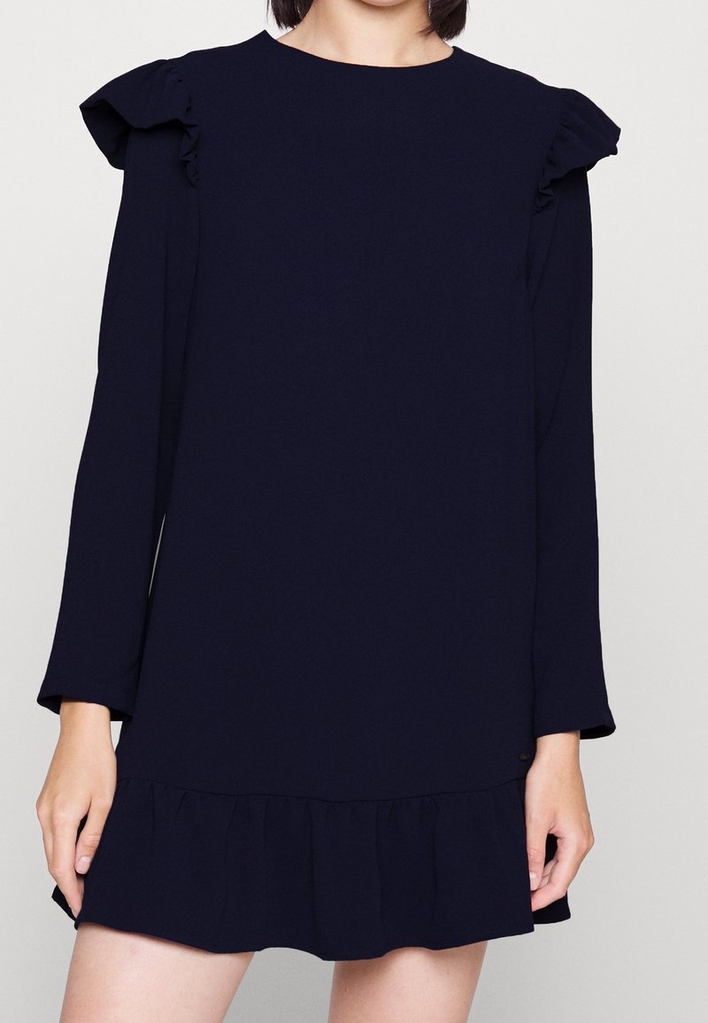 Tmavě modré šaty s dlouhými rukávy, volány na ramenou a rozšířeným tiered spodním lemem; vyrobené z hladké tkaniny s jednoduchým designem.