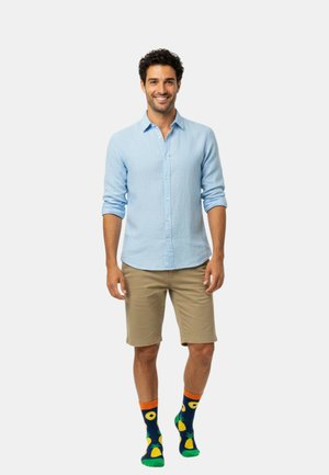 Vaaleansininen nappipaita, beiget shortsit, moniväriset sukat, joissa ananaksia ja avokadoja; rento istuvuus, pitkähihaiset ja näkyvät napit.