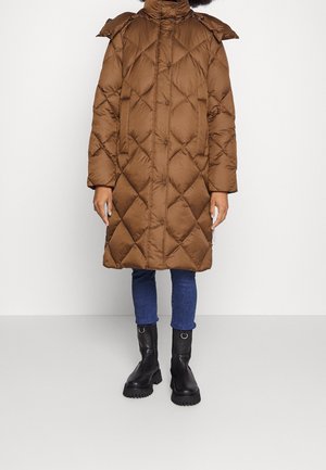 Person iført en lang brun quiltet frakke med trykknapper, blå jeans og sorte chunky støvler, stående mod en ensfarvet hvid baggrund.