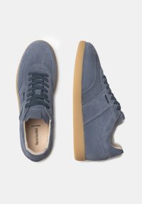 Zapatillas de gamuza azul con perfil bajo, que cuentan con una suela de goma, acentos texturizados y cordones azul oscuros. Diseño limpio con un minimalismo en la marca.