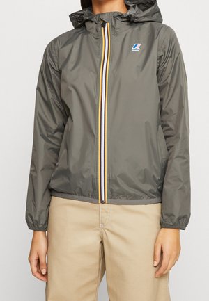 Persona con cortavientos gris con capucha, cremallera a rayas y logo azul de K-Way, combinado con pantalones beige.