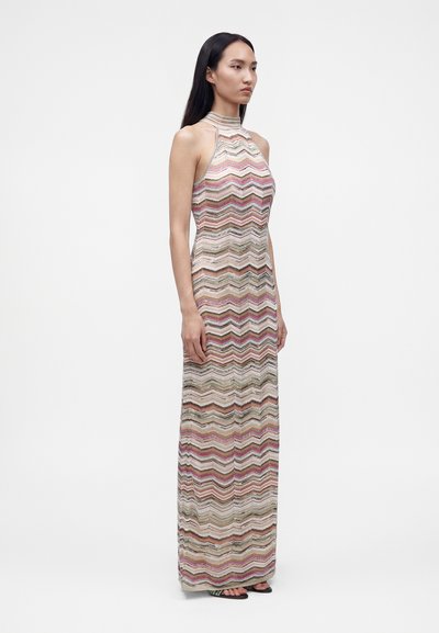 Femme debout portant une robe sans manches, col montant, longue jusqu'au sol avec des motifs zigzags multicolores et des sandales à bout ouvert.