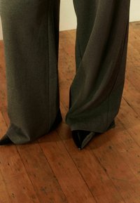 Grijze wijde broek met een gladde textuur, gecombineerd met puntige zwarte gepatineerdeleren hoge hakken op een houten vloer.