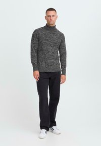 Un jeune homme portant un pull à col roulé gris à motifs, un pantalon noir et des baskets blanches avec des rayures noires, se tenant devant un fond blanc uni.