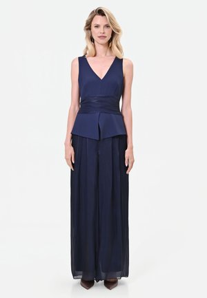 Donna in piedi che indossa un top smanicato blu navy con scollo a V, pantaloni ampi blu navy e scarpe marroni appuntite su sfondo bianco.