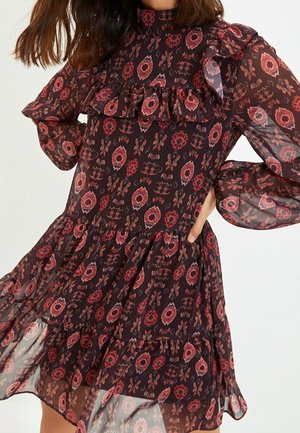 Woman wearing a long-sleeve black dress with red floral patterns, ruffles on chest and hem, posing with one hand on hip.