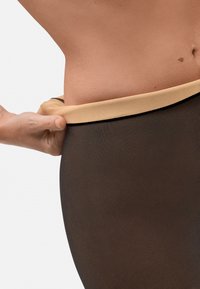Medias marrones con una textura suave y una cinturilla ancha en tono beige. Mano visible ajustando la cinturilla, resaltando el ajuste y la comodidad.