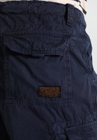 Marineblå cargo shortser laget av slitesterkt stoff, med en baklomme med et rektangulært merke og synlige sømdetaljer.
