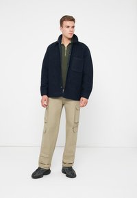 Ναυτική μπλούζα από μίγμα μαλλιού με μπροστινή τσέπη, σε συνδυασμό με ελαιόχρωμο φούτερ με φερμουάρ, μπεζ παντελόνια cargo και μαύρα παπούτσια.