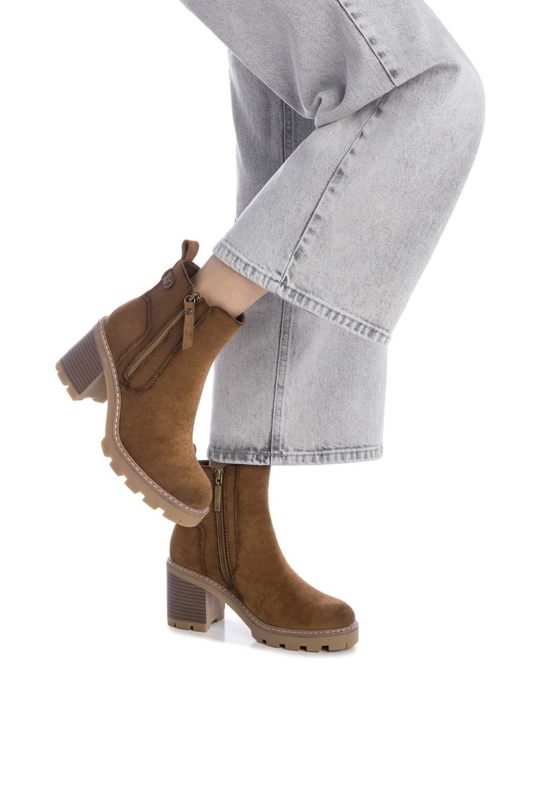 Stivaletti alla caviglia in pelle scamosciata marrone con tacco spesso, zip laterale e suola in gomma. Indossati con jeans a gamba larga grigio chiaro.