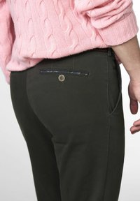 Personne portant un pantalon vert foncé avec un bouton de poche arrière et un pull en tricot rose, la main détendue sur le côté.