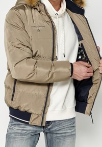 Homme portant une doudoune beige avec une capuche en fourrure ouvre sa veste pour révéler une poche intérieure noire, un sweat à capuche blanc et un jean bleu.