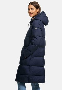 Navy blue puffer coat with a hood, featuring a quilted design, elastic drawcord, and logo patch on the sleeve. Soft, warm material.