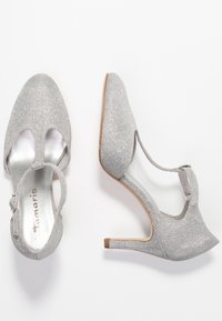 Stříbrné třpytivé lodičky s otevřenou špičkou, designem T-pásku a úzkým stiletto podpatkem. Hladká metalická podšívka a detail pásku na kotníku.