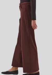 Persoon in donkerbruine wijde broek, zwarte lange mouwen top en zwarte platte schoenen, die zijwaarts staat tegen een effen achtergrond.