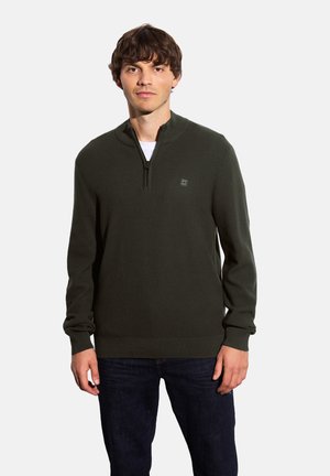 Dunkelgrüner Pullover mit Reißverschlusskragen, gerippter Textur und subtiler Logodetail auf der Brust. Trägt man über einem weißen Hemd.